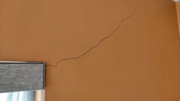 A hairline stress crack running diagonally across a painted interior wall from the head of a window casing up toward the wall-and-ceiling corner