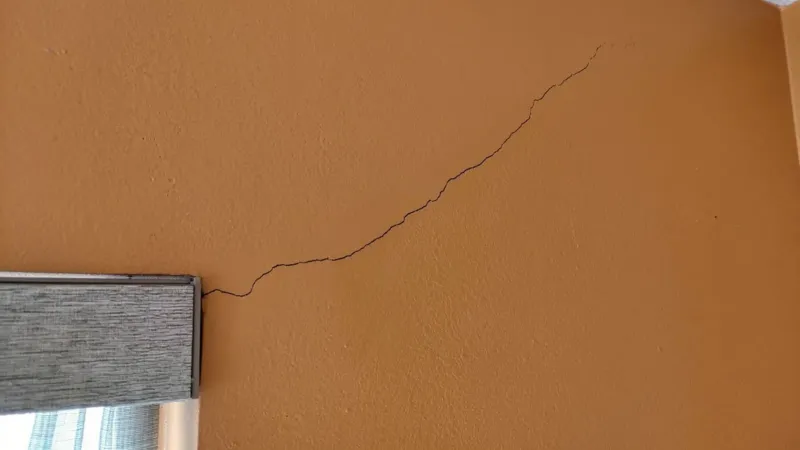A hairline stress crack running diagonally across a painted interior wall from the head of a window casing up toward the wall-and-ceiling corner