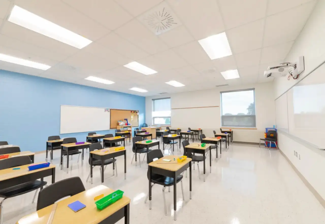 Bright school classroom with a blue accent wall of finished drywall, a suspended ceiling with recessed troffer lights, whiteboards, and rows of student desks on a tile floor