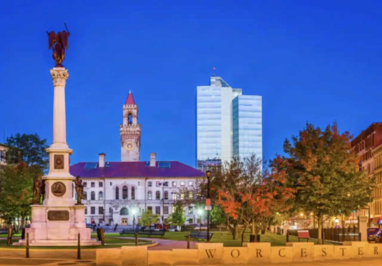 Worcester Common at dusk — the Soldiers' and Sailors' Monument, the City Hall clock tower, and the downtown glass tower rising behind, with the WORCESTER inscription along the common wall