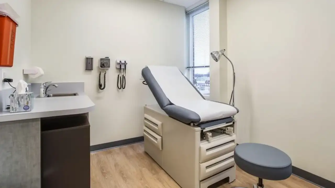 A medical exam room with an exam table against a broad plain wall, a sink counter and wall-mounted diagnostic instruments